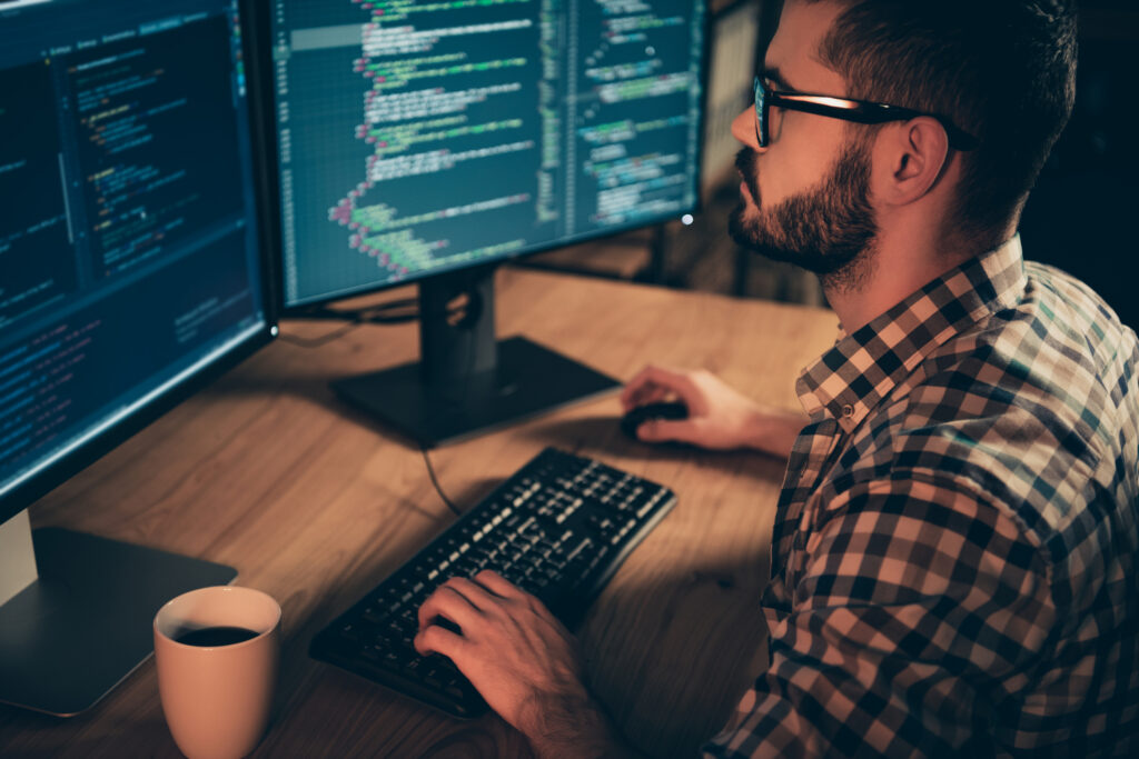 Close up side profile photo handsome he him his guy linux windows cross platform coder typing php css keyboard development outsource IT monitors table office agency wear specs formalwear plaid shirt